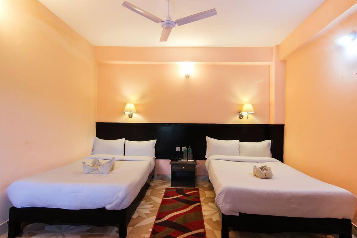 Quarto de hotel com duas camas, um ventilador de teto, luminárias laterais e uma mesa de cabeceira com abajur.