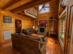 A log cabin interior with a fireplace, a television, a ceiling fan, and a wooden staircase.