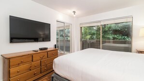 Un dormitorio con una cama grande, un aparador de madera, un televisor de pantalla plana y un ventanal corredero que da a un balcón con vistas a la vegetación.