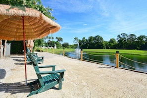 A beach area with a thatched umbrella, green chairs, and a water feature.