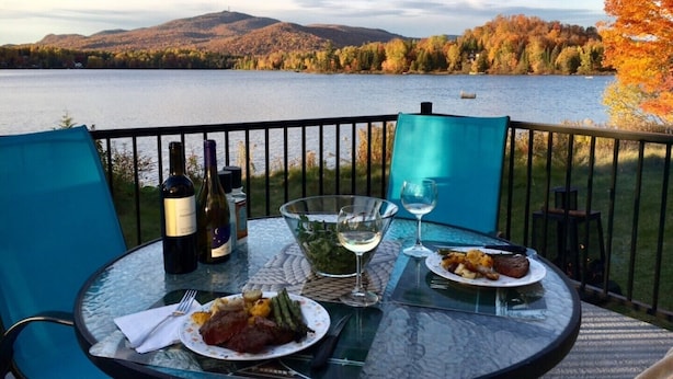 A table set with wine, glasses, and plates of food, overlooking a lake with autumn foliage and mountains in the background. - Lakefront Chalet Mont Tremblant - Spectacular view - WIFI (CITQ 294661) (Lac-Supérieur)
