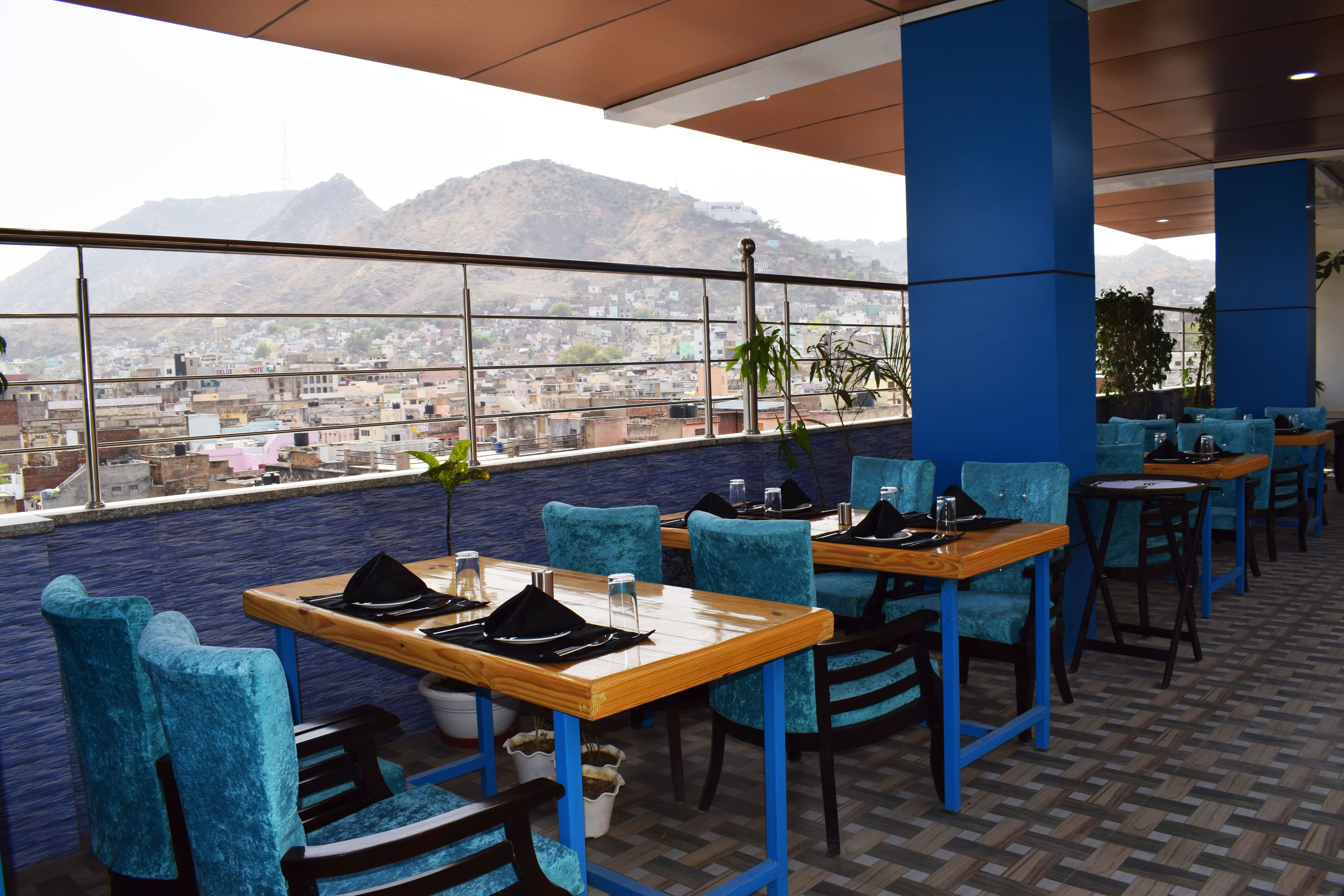 A restaurant with blue chairs and wooden tables, a city view, and mountains in the background.
