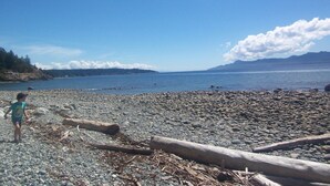 A rocky beach with a child playing, a calm sea, and mountains in the distance. - Private waterfront cottage on Malaspina Strait with easy access to town/trails (Powell River)