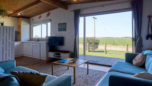 A spacious living area with a large window, a kitchen with white cabinets, a flat-screen TV, and a cozy blue sofa. - Far View - Pets,  Linen,  Wi-Fi, Netflix, Caravan, EV charger (Venus Bay)