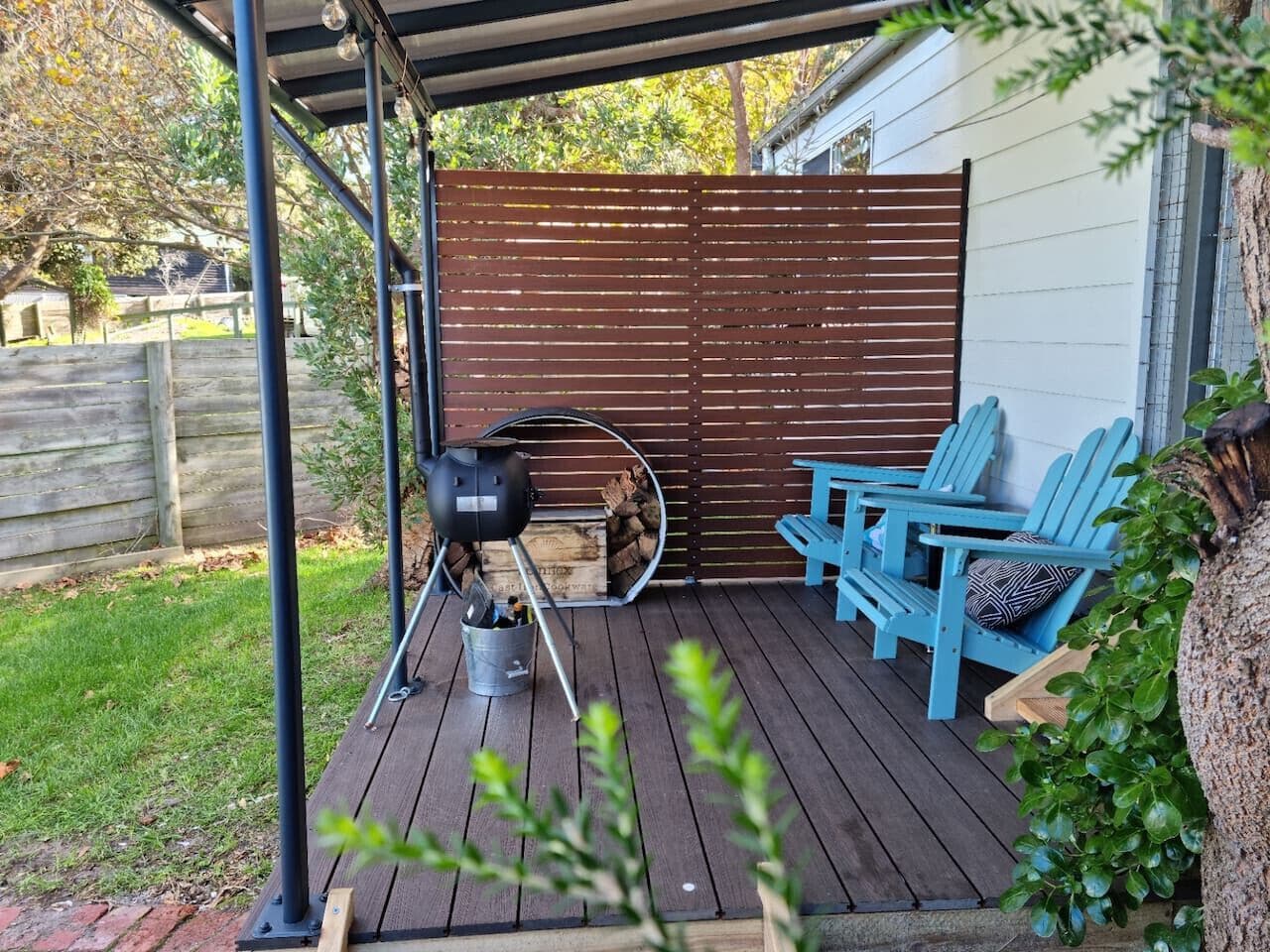 A covered patio with a grill, blue Adirondack chairs, and a wooden fence.