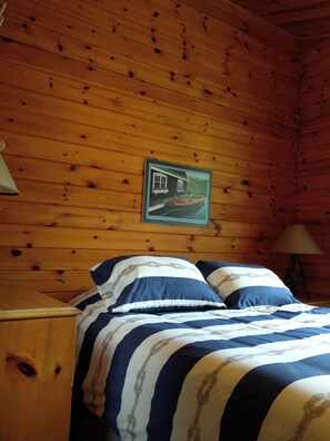 A wooden cabin room with a bed, a nightstand, a lamp, and a framed picture on the wall.
