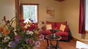 A cozy living room with a red sofa, a small round table with a flower vase, and a window with a view of trees.