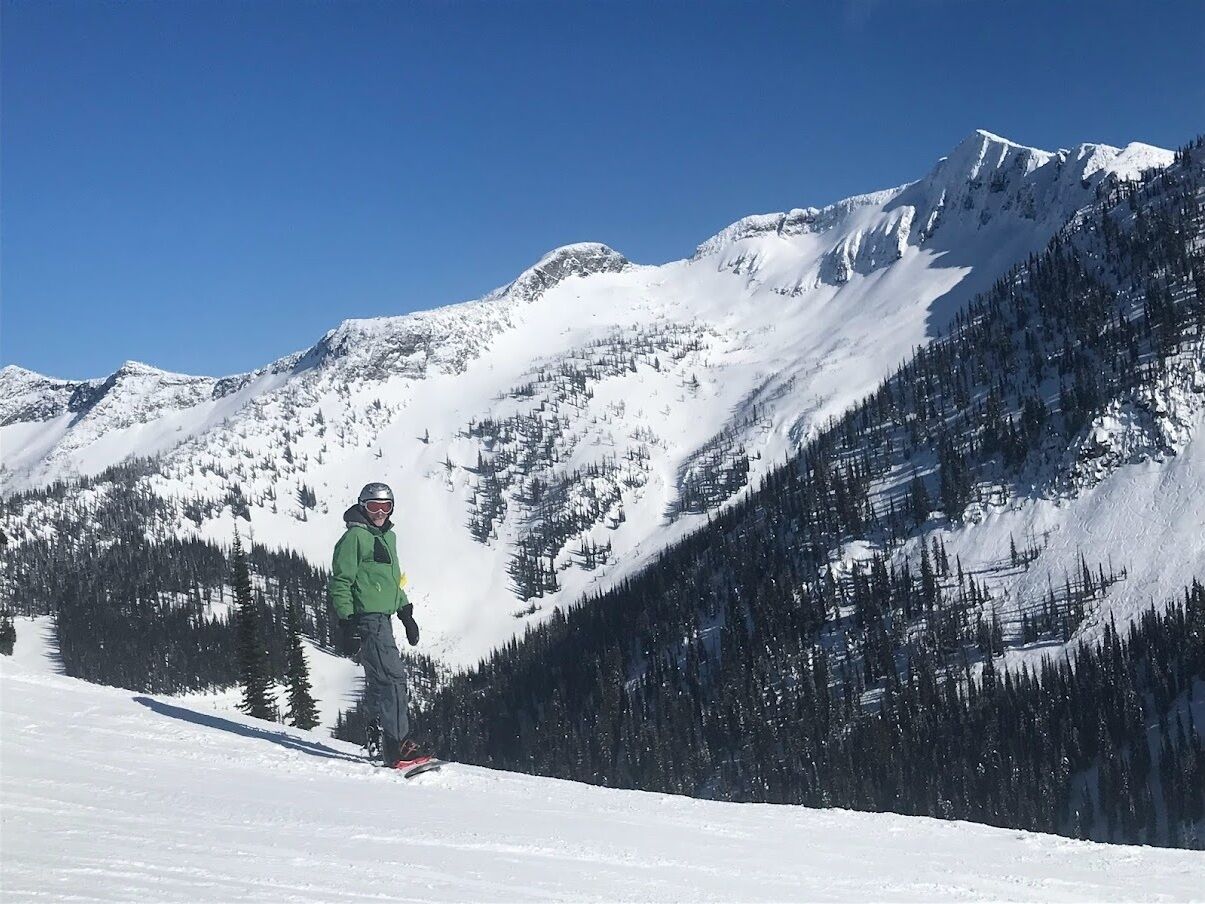 Een persoon snowboardend op een besneeuwde berghelling, met dichte bossen en met sneeuw bedekte toppen op de achtergrond.
