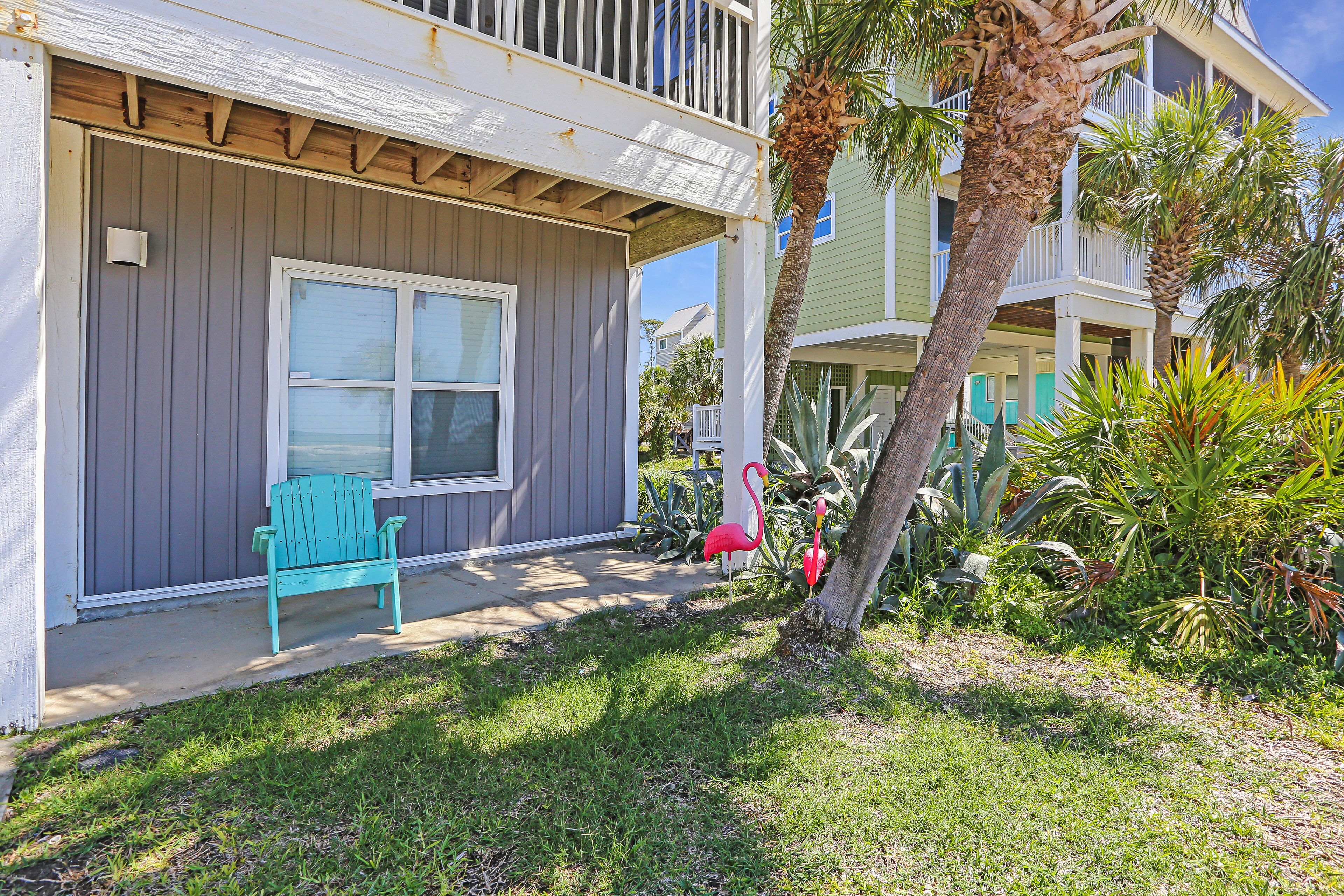 A beachfront property with a covered porch, a turquoise Adirondack chair, and a small garden with a flamingo statue.