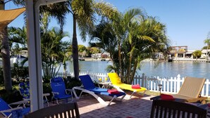 Un espace aménagé au bord de la piscine, avec des chaises longues, une table avec des assiettes et des tasses, et une vue sur un plan d’eau bordé de palmiers et délimité par une clôture blanche.