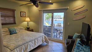 Une chambre à coucher comprenant un lit, un ventilateur de plafond, une fenêtre avec des stores, une lampe, un téléviseur et une vue sur un balcon aménagé avec du mobilier d’extérieur.