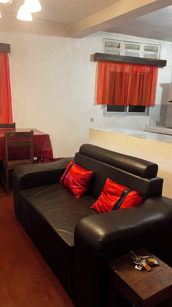 A living room with a black sofa, red cushions, a wooden table, and a window with orange curtains.