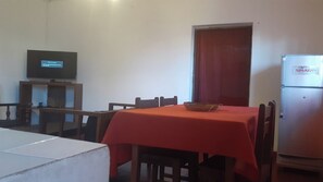 A dining area with a table covered in a red tablecloth, wooden chairs, a television on a stand, and a refrigerator. - Villa Cali Paso, Sambava (Sambava)