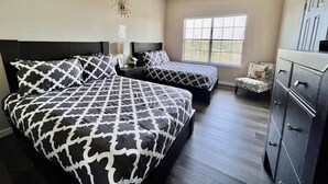 A bedroom with two beds, a ceiling fan, a window with blinds, a nightstand, a dresser, and a small chair.