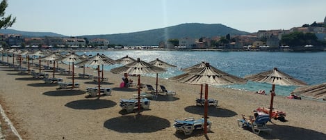 A beach with rows of sun umbrellas and lounge chairs, a clear view of the sea, and buildings in the background.