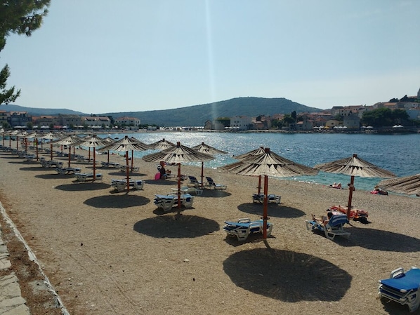 A beach with rows of sun umbrellas and lounge chairs, a clear view of the sea, and buildings in the background. - Summer cottage with breathtaking view (Zecevo Rogoznicko)