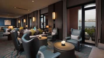 Een hotellobby met een zithoek, een groot raam met uitzicht op een waterpartij en gebouwen, en een schilderij aan de muur.