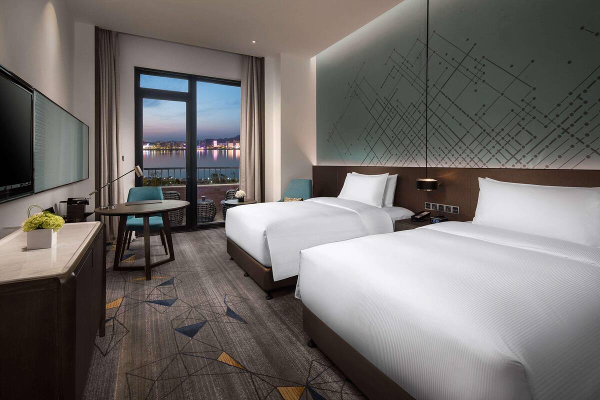 Ein Hotelzimmer mit zwei Betten, einem Schreibtisch, einem Sessel, einem Fernseher und einem großen Fenster mit Blick auf die Stadt.