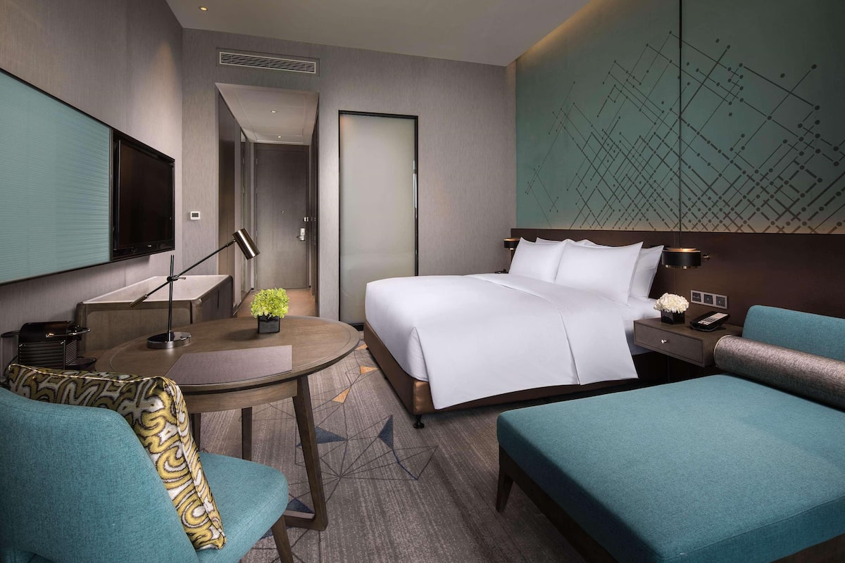 Una habitación de hotel moderna con una cama grande, un escritorio con lámpara, una pequeña mesa con planta y un área de descanso con sofá y sillón.