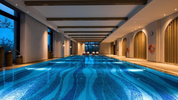 A long indoor swimming pool with a tiled floor, wooden ceiling beams, and a view of the outdoors through large windows. - Doubletree by Hilton Qingdao Oriental Movie Metrop (Qingdao)
