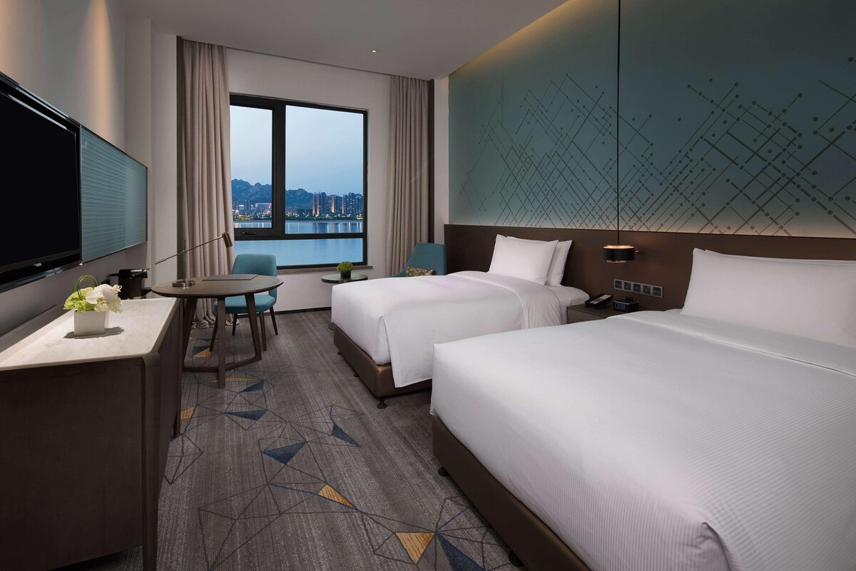 A hotel room with two beds, a desk, a TV, and a window with a city view.