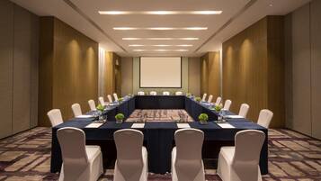 A conference room with a long table, white chairs, and a projector screen.