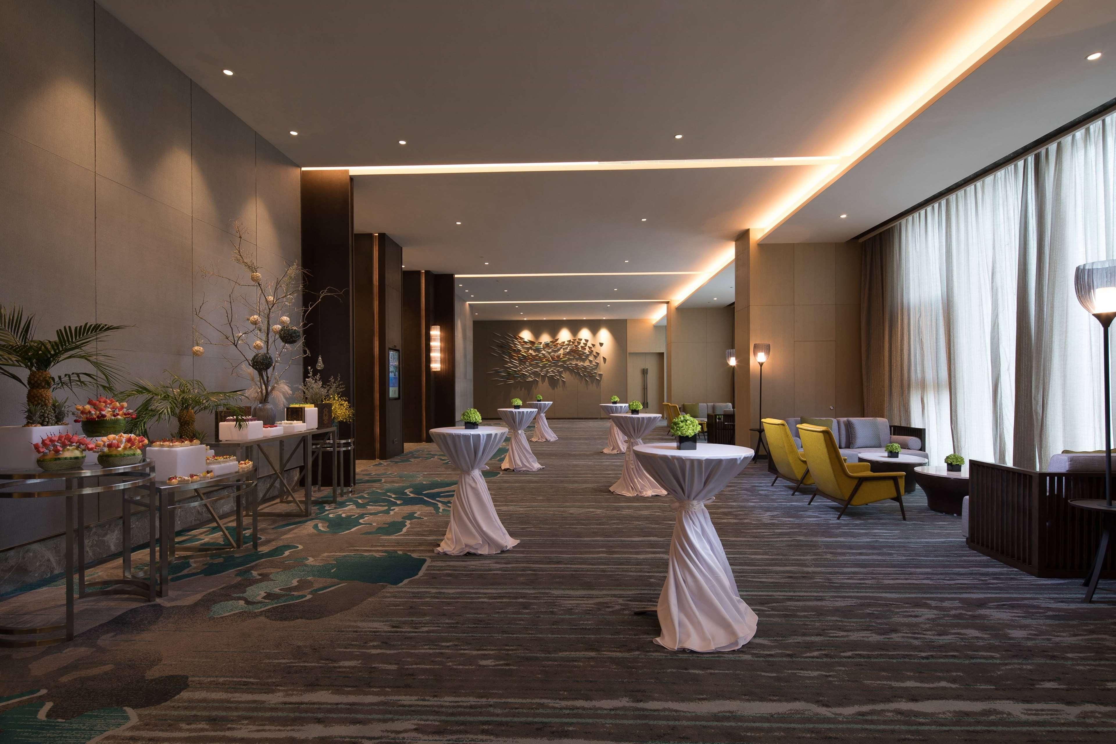 A hotel lobby with round tables, chairs, a reception desk, and a large window with curtains.