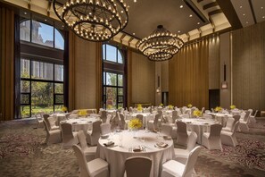 Un salón de banquetes con mesas redondas, sillas blancas, candelabros y ventanas grandes.