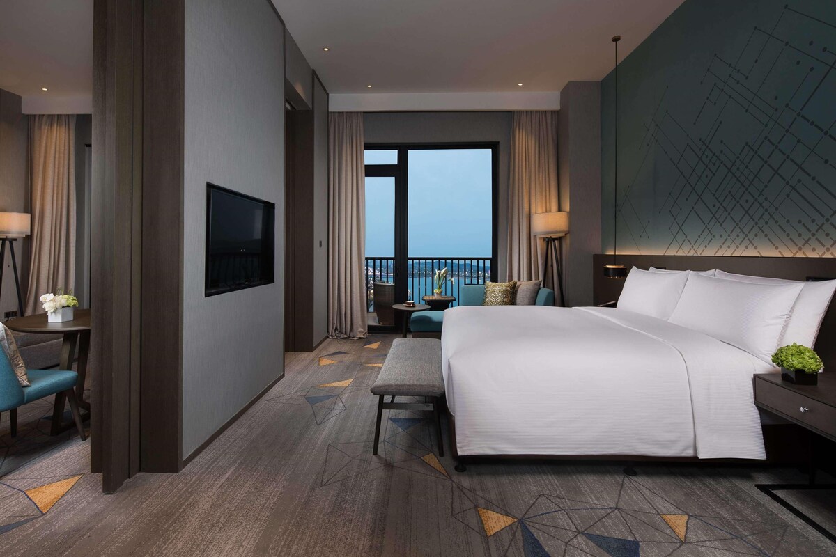 A modern hotel room with a large bed, a TV, a balcony with a view, and a desk with a chair.
