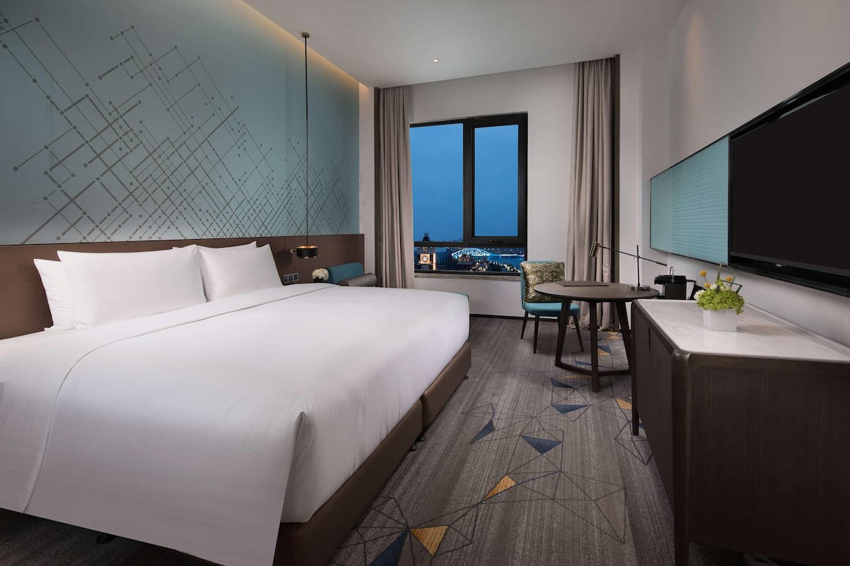 Ein Hotelzimmer mit einem großen Bett, einem Schreibtisch, einem Sessel, einem Fernseher und einem Fenster mit Blick auf die Stadt.