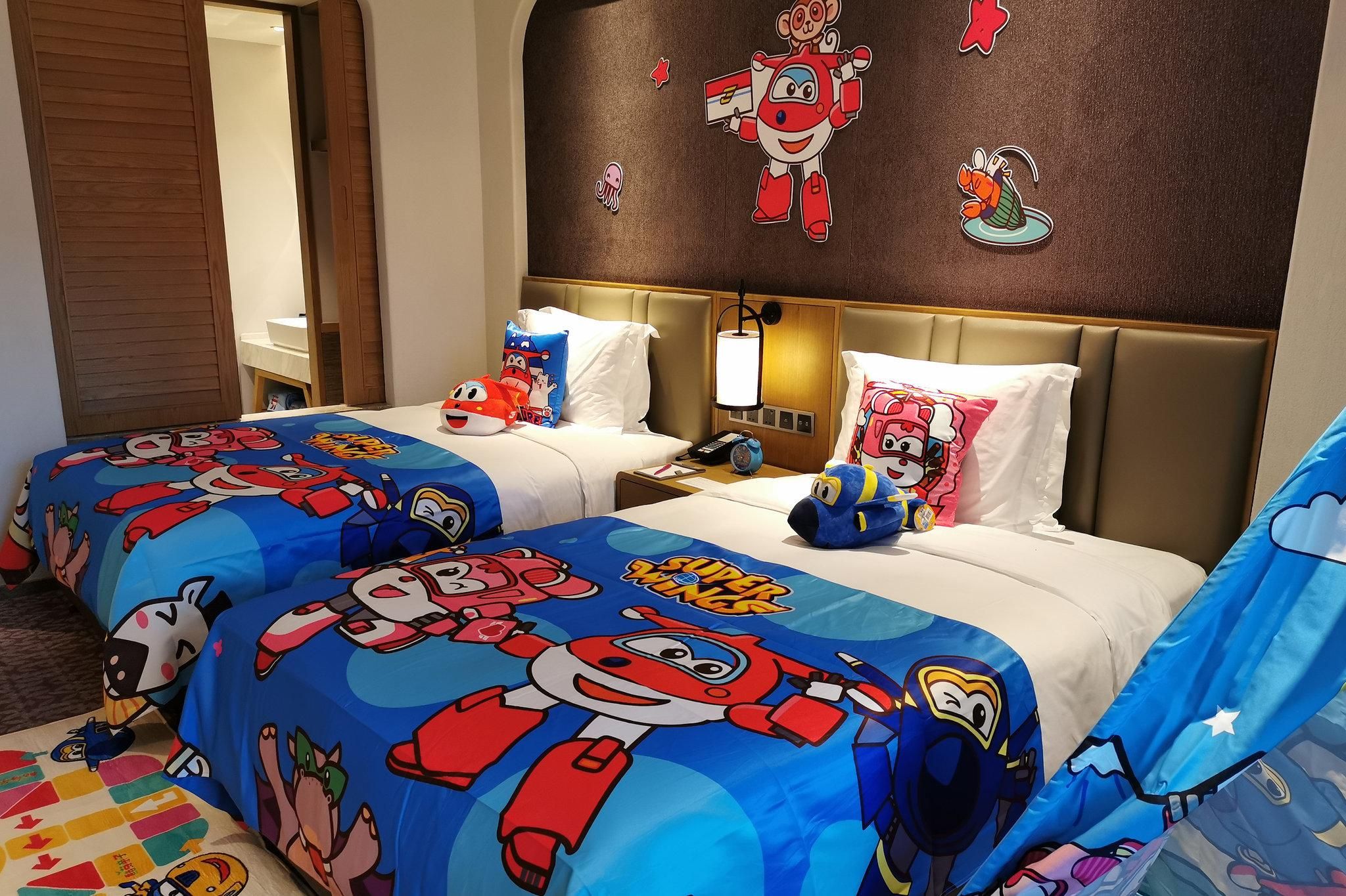 A hotel room with two beds, each covered with a Super Wings themed duvet. There are pillows with Super Wings characters, a nightstand with a lamp, and a wall with Super Wings stickers.