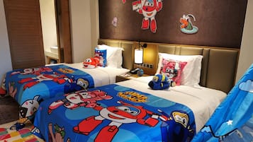 A hotel room with two beds, each covered with a Super Wings themed duvet. There are pillows with Super Wings characters, a nightstand with a lamp, and a wall with Super Wings stickers.