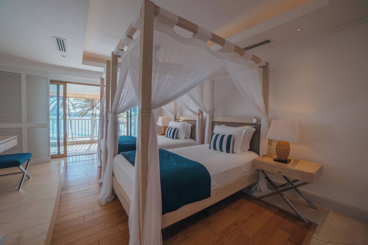 Una cama con dosel sobre cuatro pilares con vista a palmeras, suelo de madera y balcón.