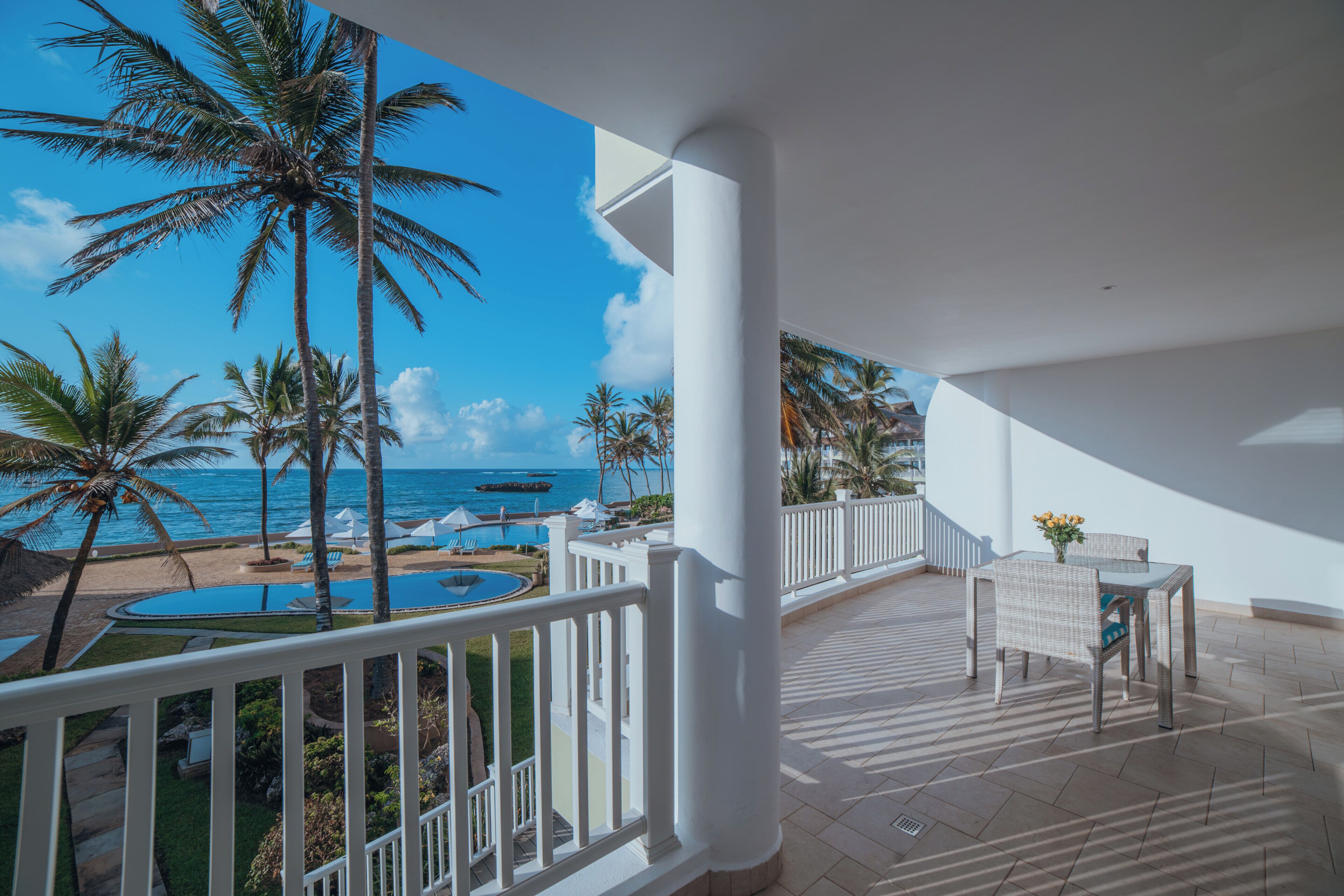 A balcony with a view of the ocean and palm trees, a pool, and outdoor furniture.