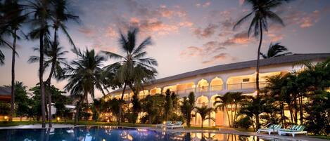 A hotel with a swimming pool, palm trees, and a sunset sky.