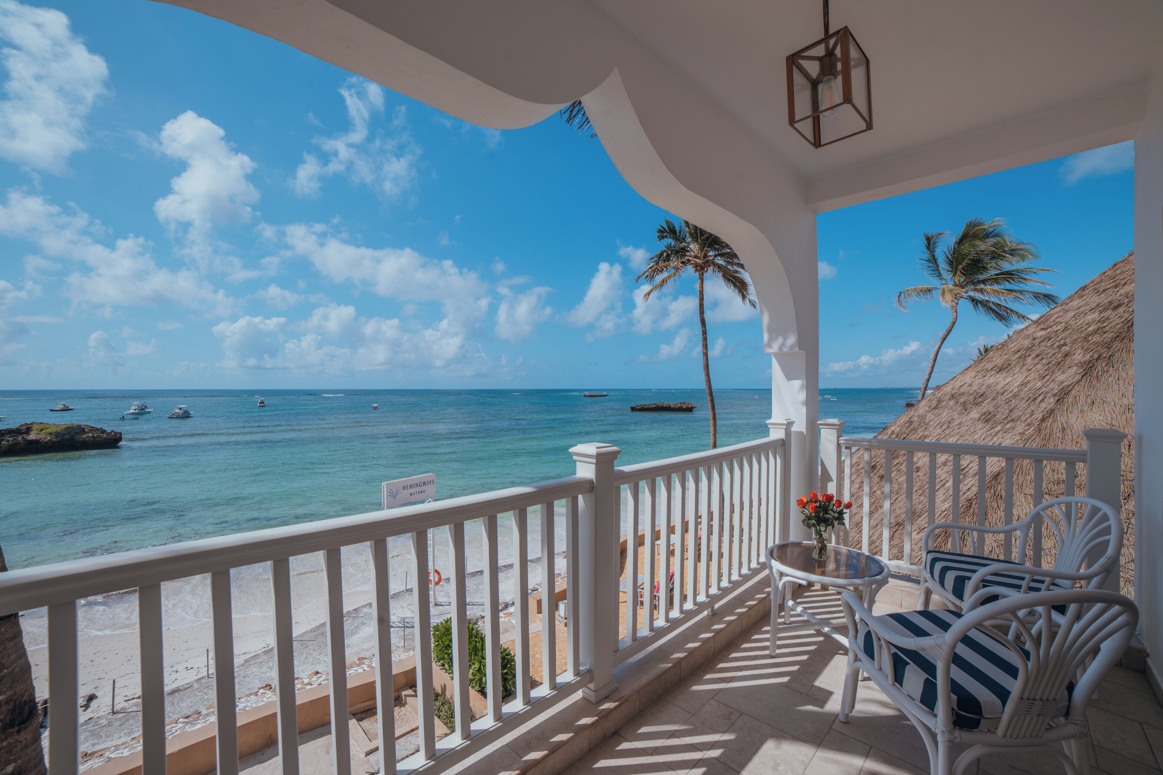 A balcony with white railings, wicker furniture, and a view of the sea with boats and palm trees.