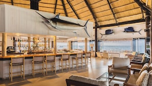 Zona de bar con vista al mar, mobiliario de madera y techo de paja.