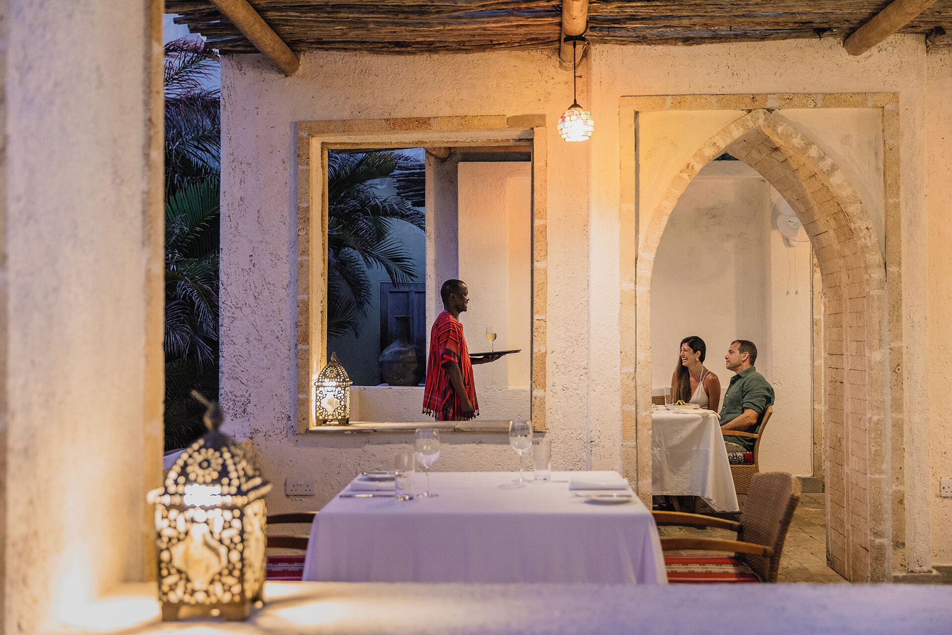 An outdoor dining area with a table set for two, a waiter carrying a tray, and a window with a view of palm trees.