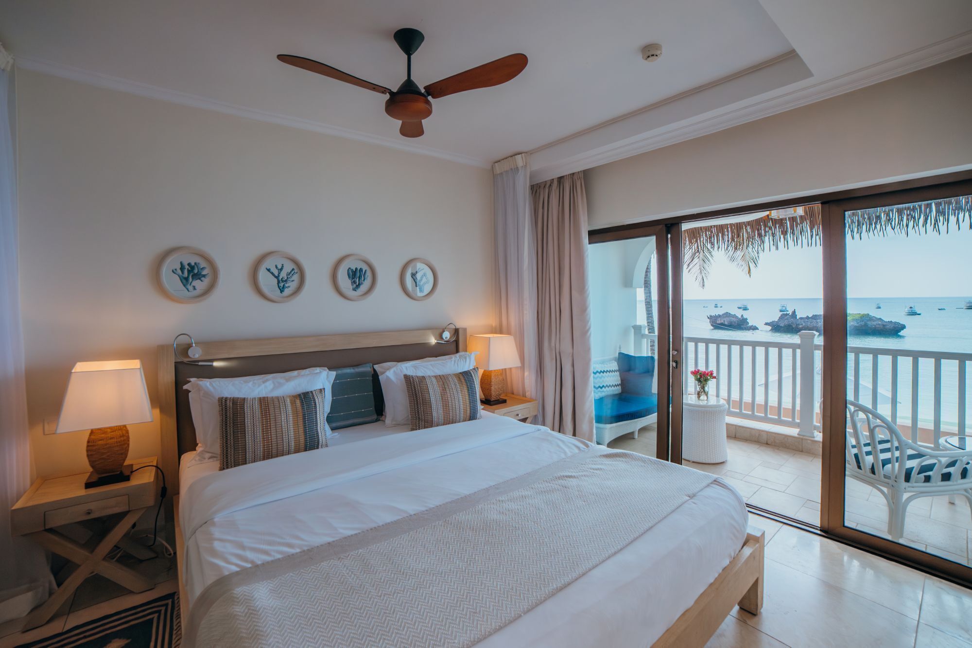 A hotel room with a bed, a ceiling fan, a nightstand, a lamp, a view of the sea, and a balcony with chairs.