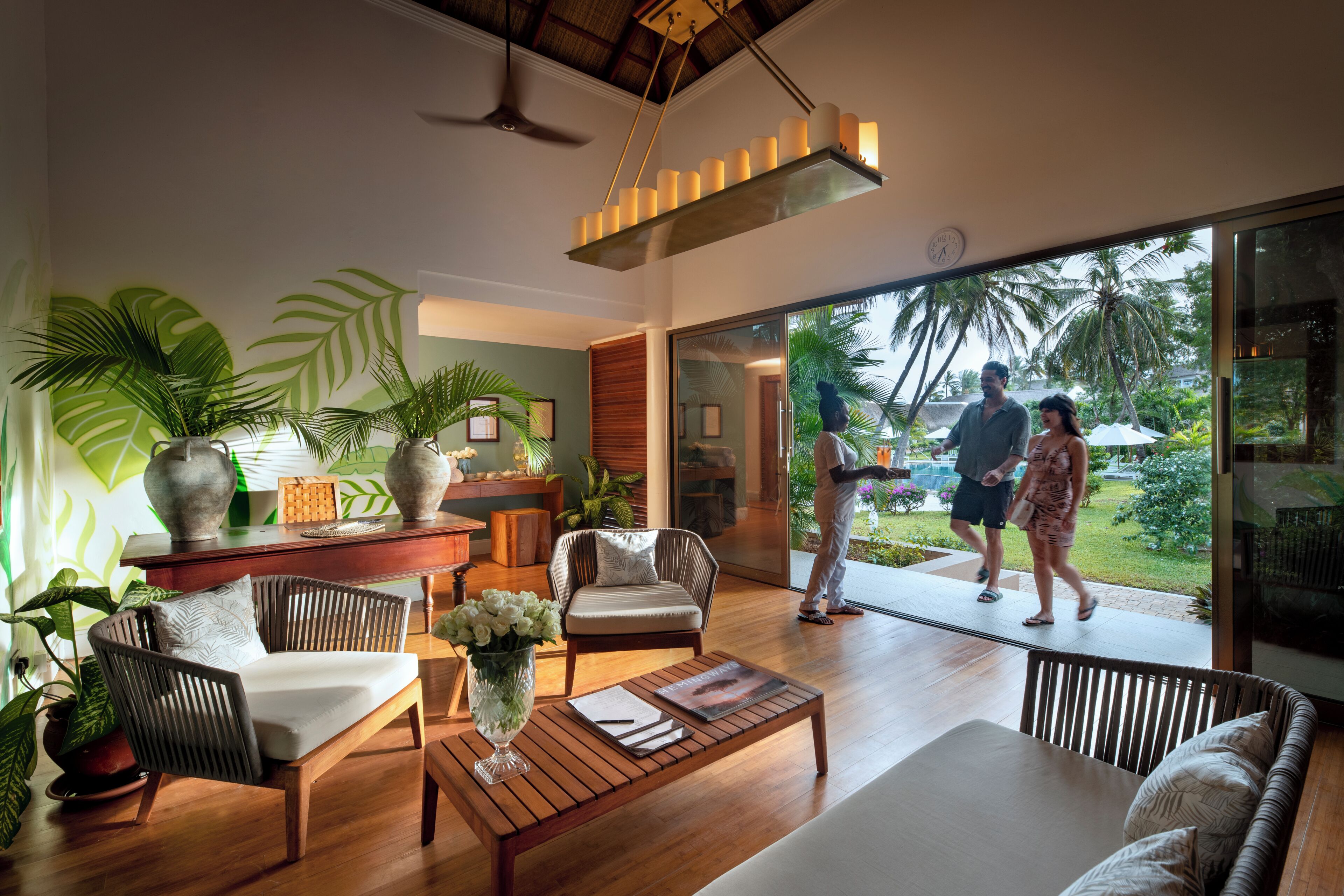 A modern living room with a wooden coffee table, a sofa, and a chair. There is a large window with a view of palm trees and a balcony.