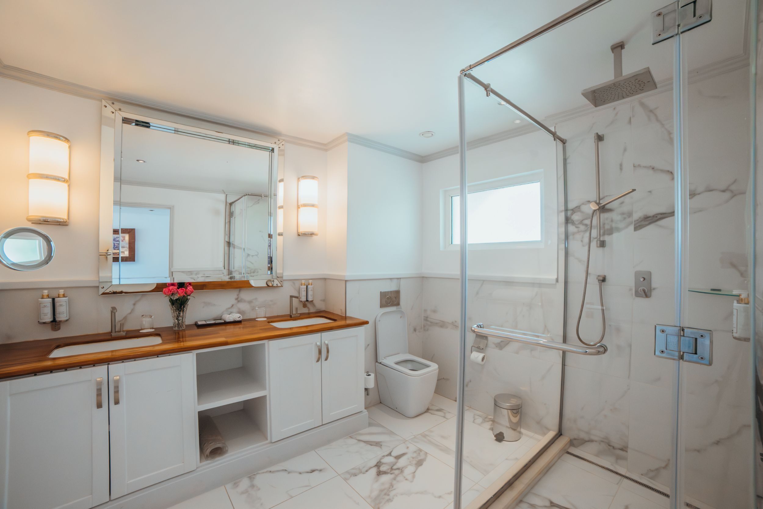 A modern bathroom with a glass shower enclosure, a wooden vanity, a white toilet, and marble flooring.