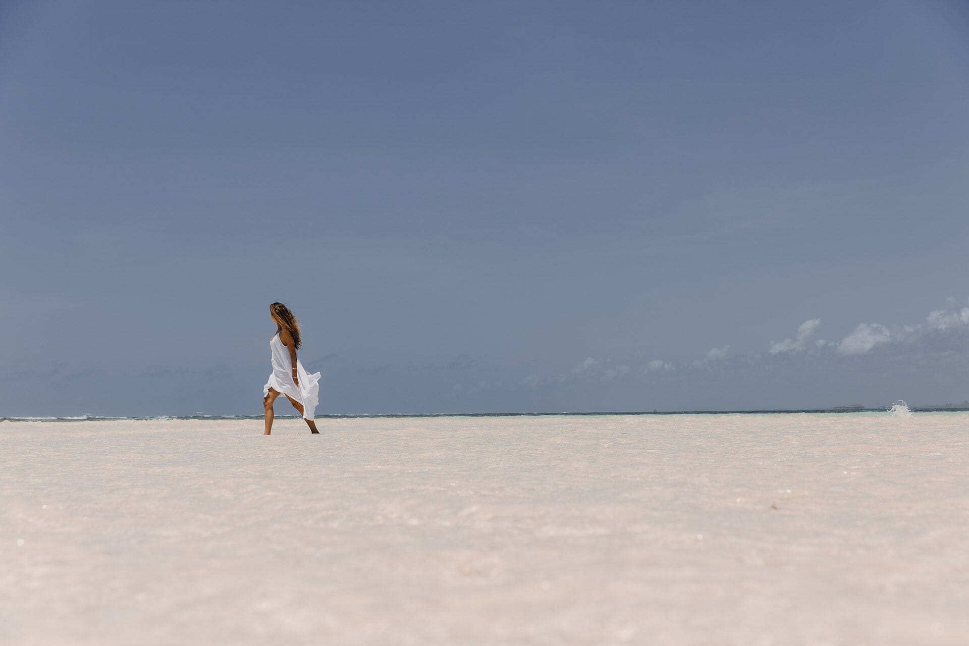 A person in a white dress walking on a beach with a clear sky.