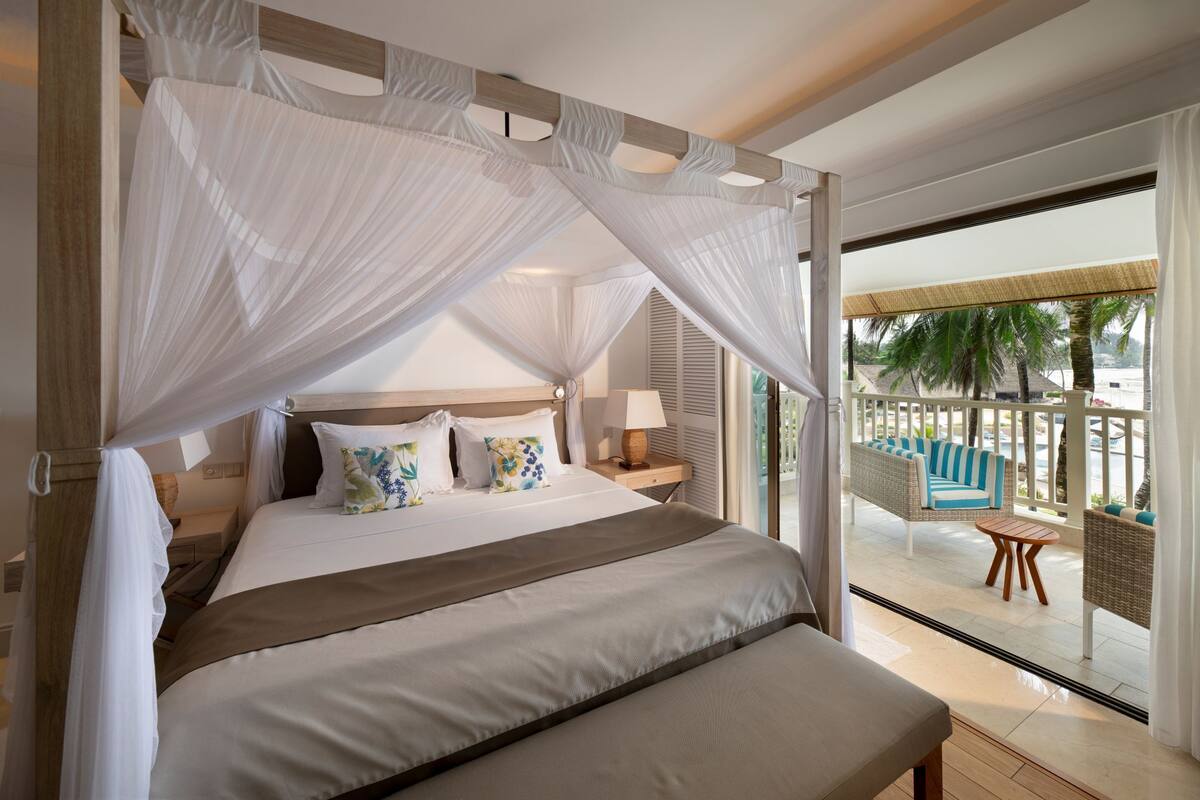 Un dormitorio con cama a dosel, un balcón con mobiliario de mimbre y vistas a palmeras.