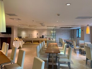 A modern dining area with long tables and cushioned chairs, a kitchen counter, and a large window providing natural light. - JI Hotel (Shenzhen)