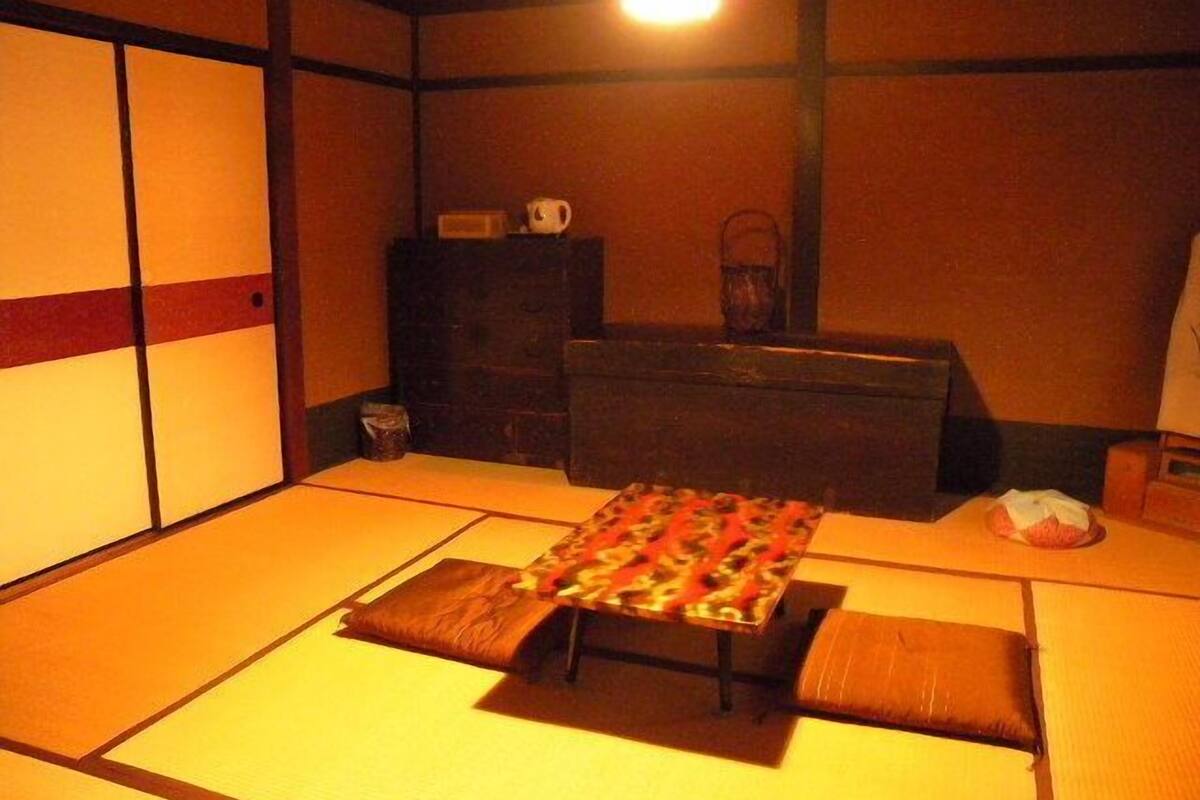 Een kamer met tatami matten, een lage tafel, kussens, een klein kastje en een hanglamp.