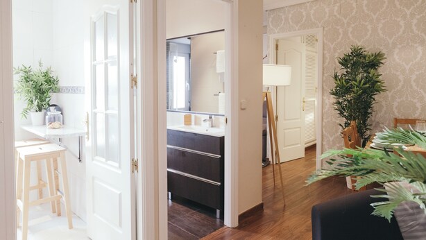 Un baño moderno con un lavabo oscuro, azulejos blancos y un espejo. Hay un taburete de madera y una planta en maceta cerca de la entrada.
