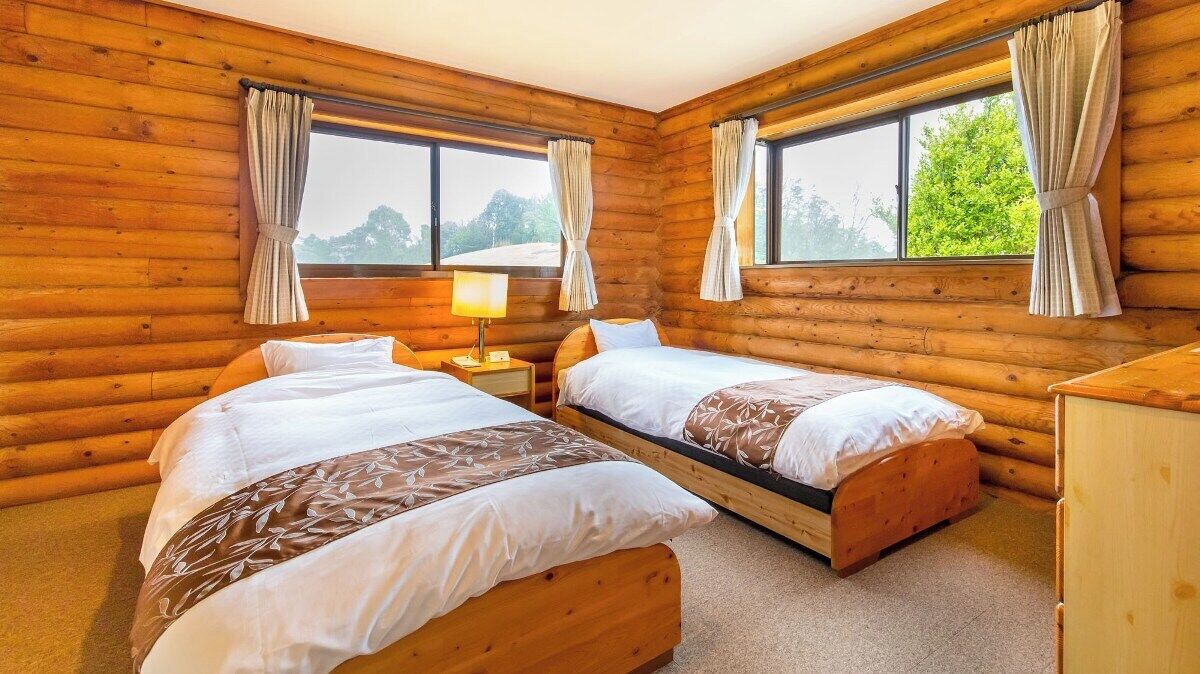 A log cabin room with two beds, a nightstand, a lamp, and large windows with curtains.