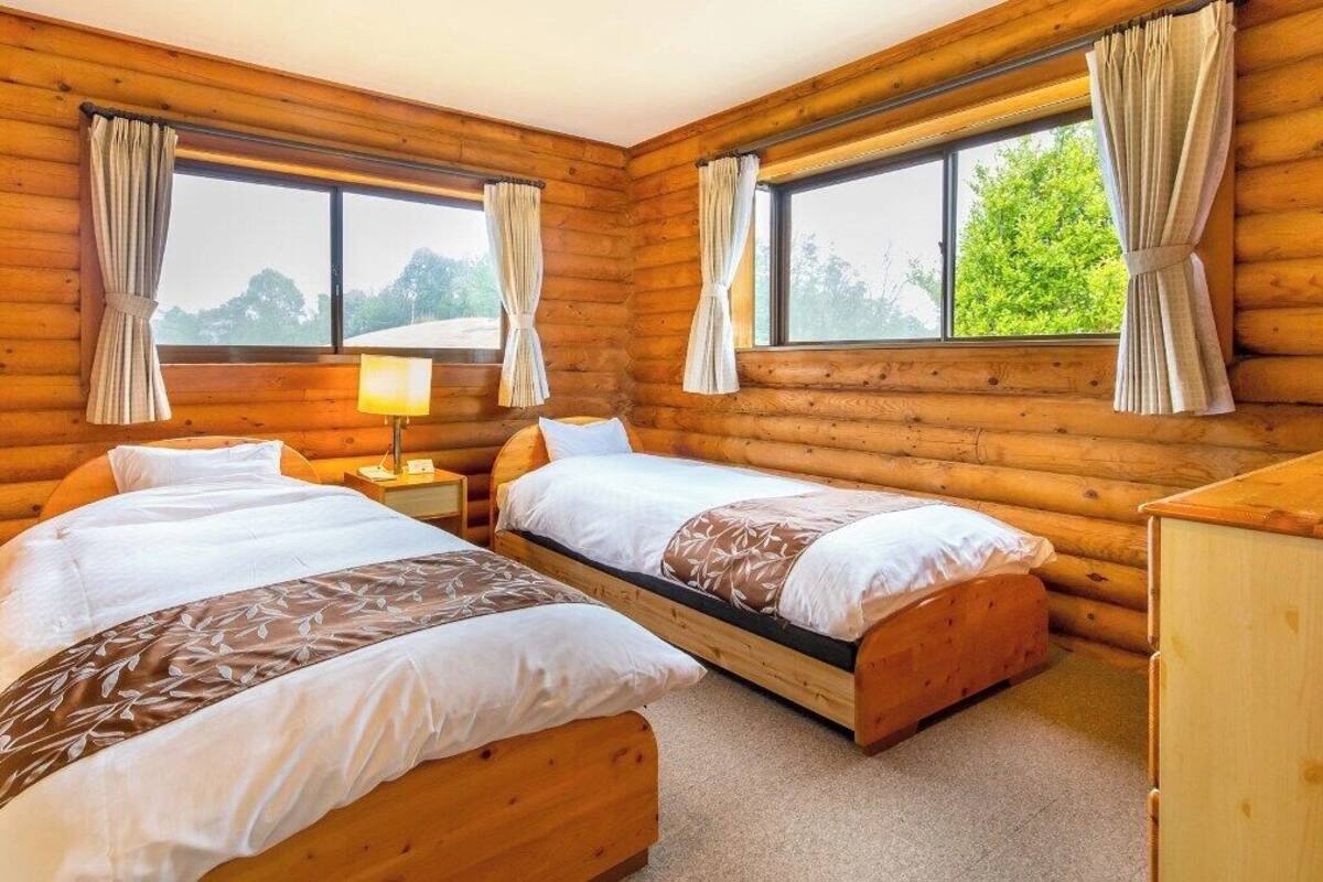 A log cabin room with two beds, a nightstand, a lamp, and large windows with curtains.