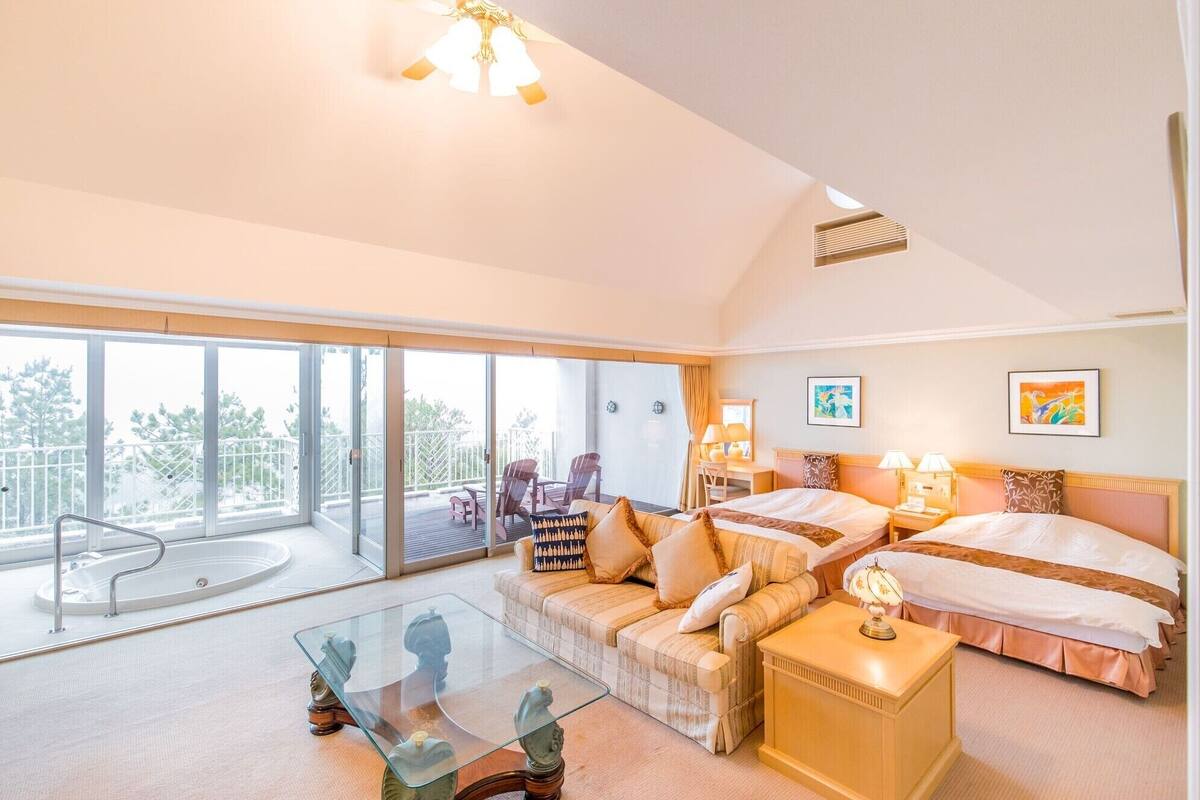 A hotel room with a balcony, a sofa, a glass coffee table, two beds, and artwork on the walls.