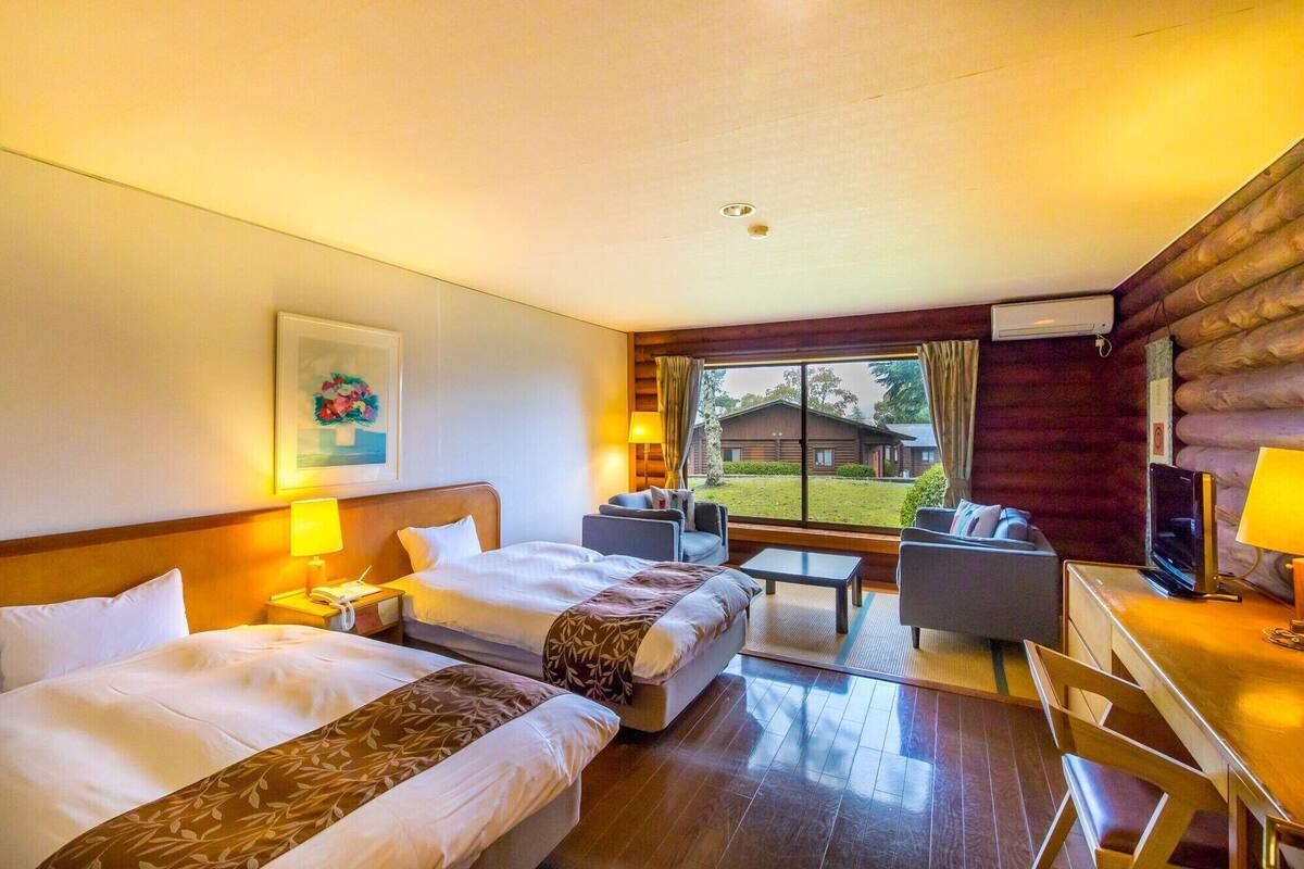 A hotel room with two beds, a desk, a chair, and a view of a cabin.